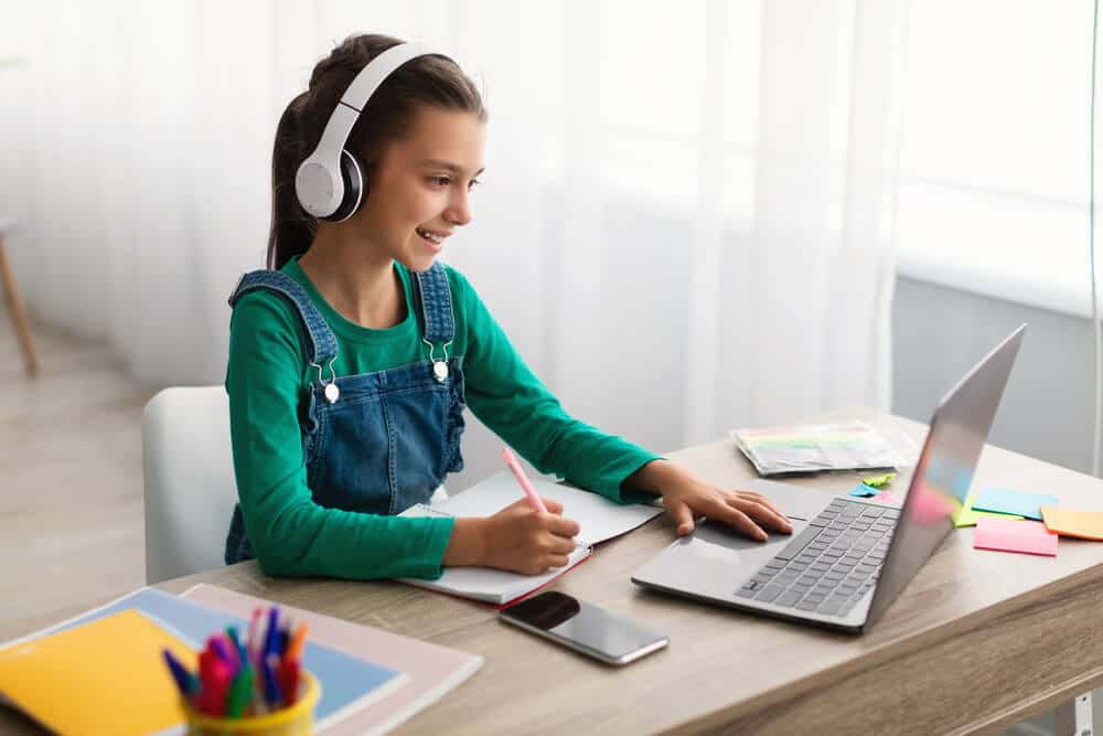 girl sitting at desk using laptop and writing on a pad